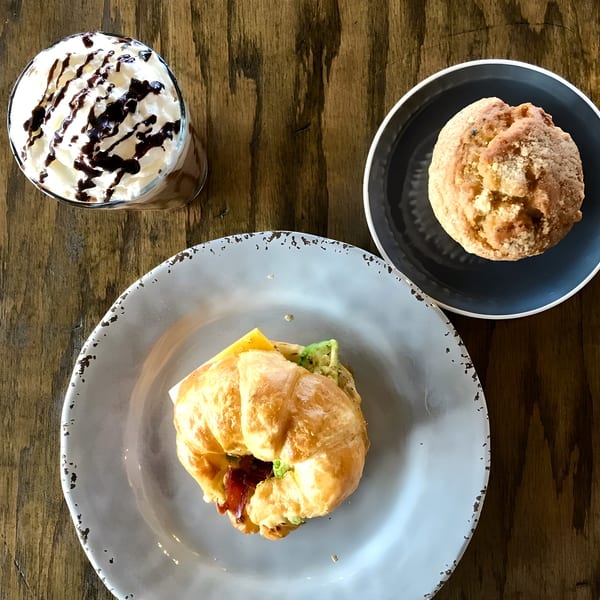 A Crave Coffee & Tea croissant sandwich, muffin, and iced coffee on a wooden table.