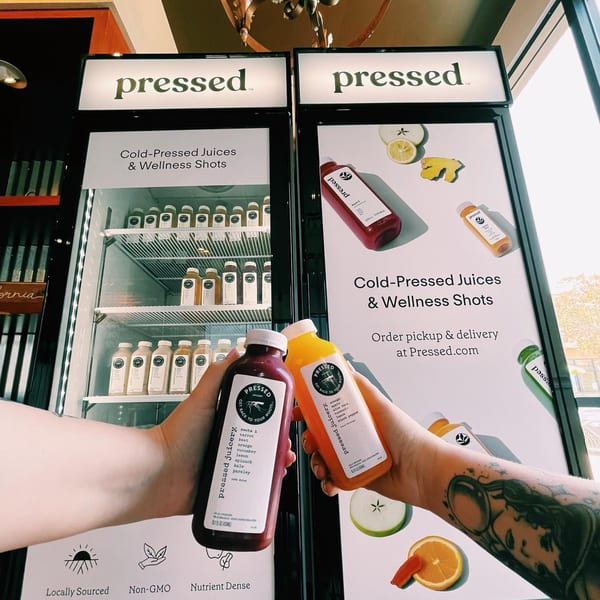 Hands holding two Pressed Juicery bottles in front of a display case and promotional sign.