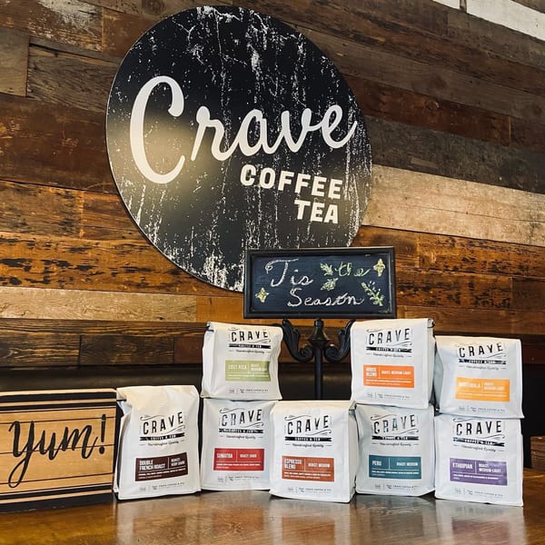 Bags of Crave Coffee & Tea beans on a wooden counter with a "Crave" sign in the background.