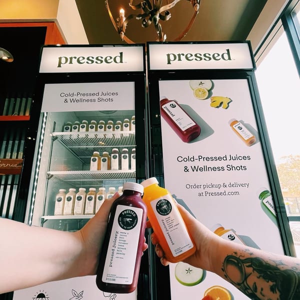Two hands holding Pressed Juicery bottles in front of a display of cold-pressed juices.
