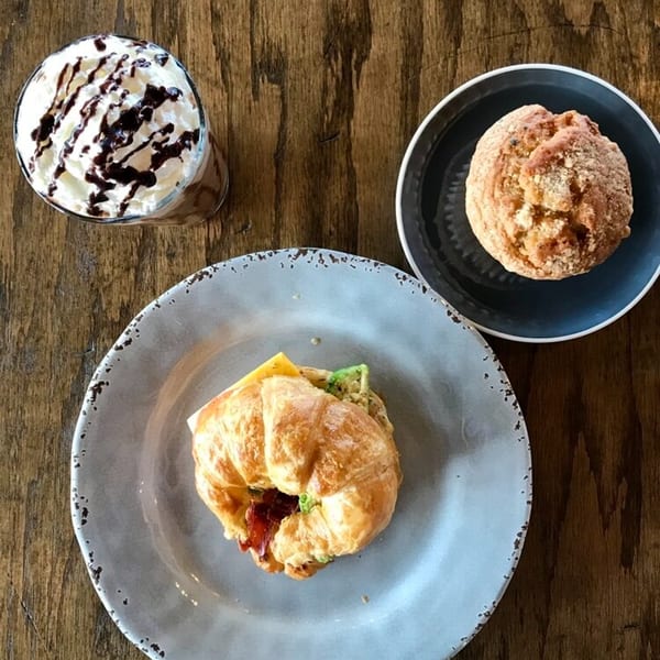 Overhead view of a croissant sandwich, muffin, and iced coffee with whipped cream on a wooden table.